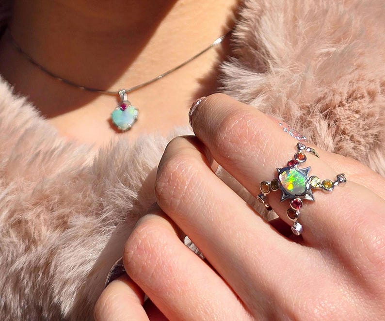 May include: A silver ring featuring a central iridescent opal surrounded by small, colourful round gemstones, paired with a matching silver necklace pendant that also displays a single opal and a small red gemstone accent.