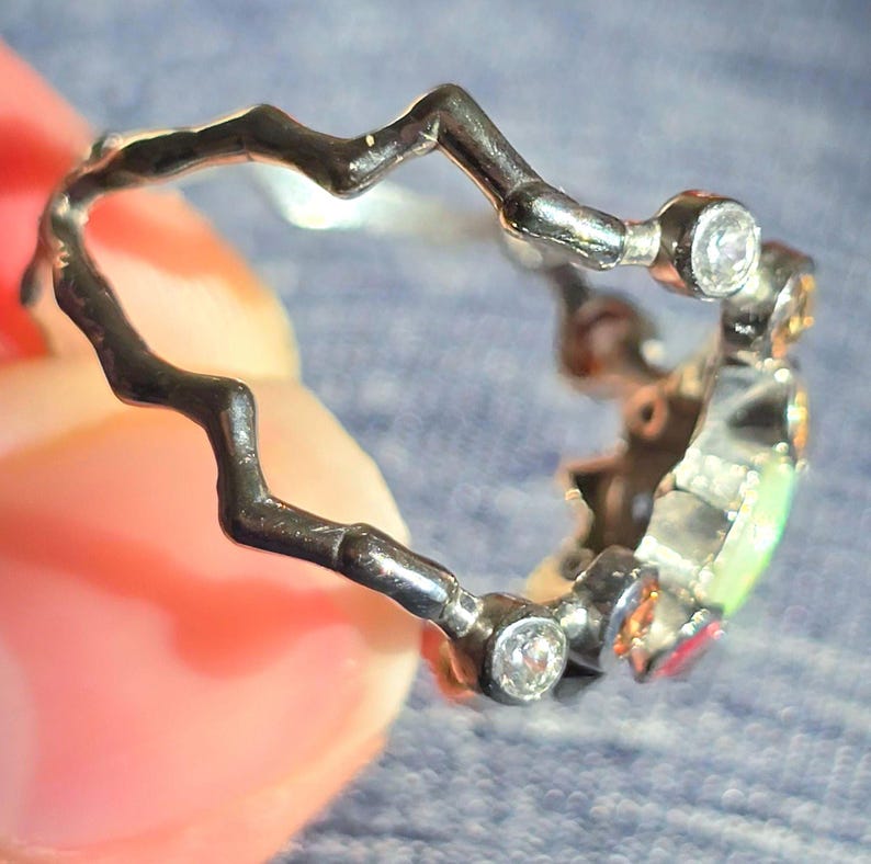 May include: A dark metal ring featuring a jagged, zigzag-shaped band. The ring is accented with several small, clear round stones and a central, iridescent oval-shaped stone that displays green and orange hues.