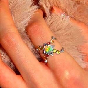 May include: A silver-toned ring featuring a central oval-shaped iridescent opal stone set within a star-shaped frame, accented by smaller round multi-coloured gemstones along the split band design.