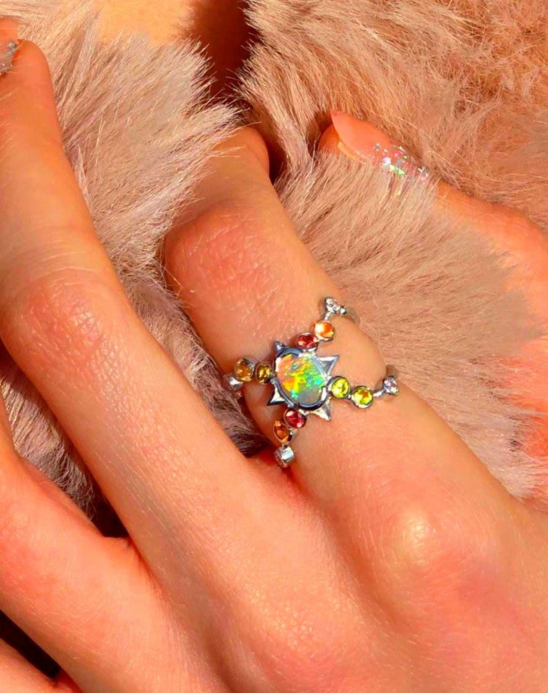 May include: A silver-toned ring featuring a central oval-shaped iridescent opal stone set within a star-shaped frame, accented by smaller round multi-coloured gemstones along the split band design.