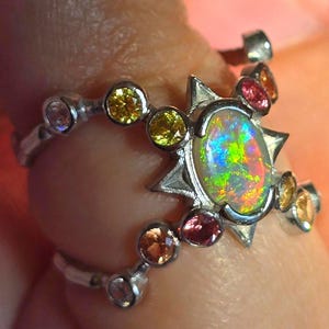 May include: A silver ring featuring a central oval opal with a vibrant play of colour, surrounded by a star-shaped setting. The band is accented with multiple small, round, multi-coloured gemstones including yellow, pink, and orange stones.