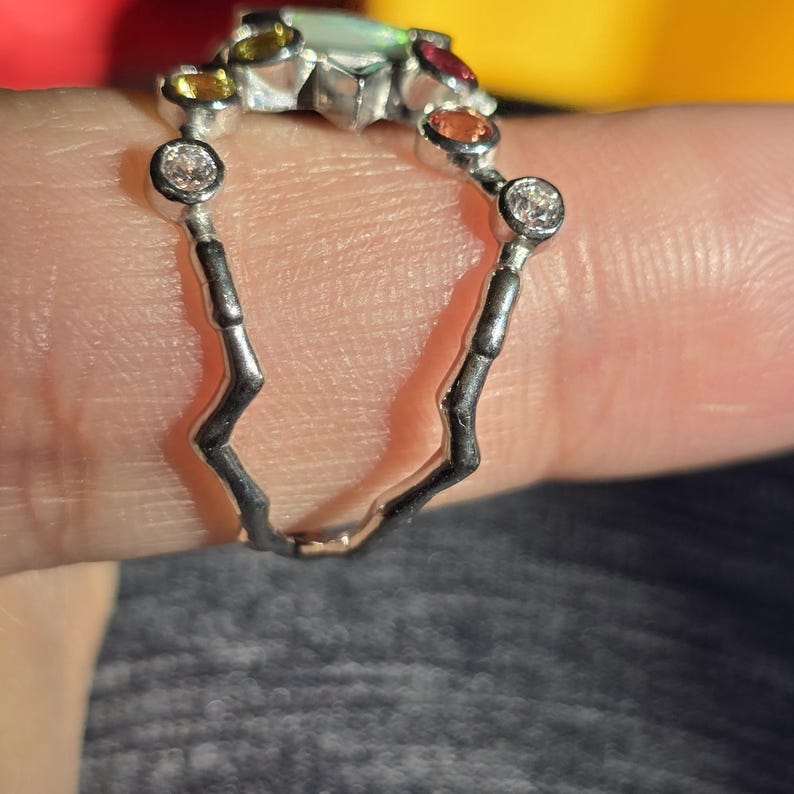 May include: A silver ring featuring a jagged, textured band and a top row of multi-coloured gemstones, including clear, yellow, orange, and red stones, set in individual circular bezels along the upper arc of the metal structure.