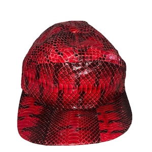 Puede incluir: Gorra de béisbol roja con un patrón de piel de serpiente. La gorra tiene una visera curva y una corona redondeada. El patrón de piel de serpiente es una mezcla de rojo y negro, creando una apariencia texturizada. Es un accesorio elegante.