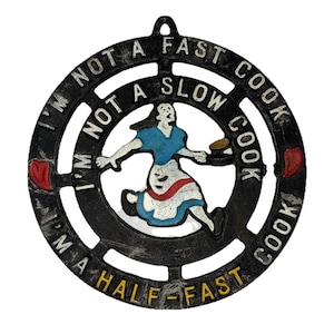Puede incluir: Salvamanteles de hierro fundido negro con el texto "I'M NOT A FAST COOK, I'M NOT A SLOW COOK, I'M A HALF-FAST COOK." El salvamanteles presenta a una mujer con un vestido azul y un delantal rojo que lleva un plato. Dos formas de corazón rojo en los lados.