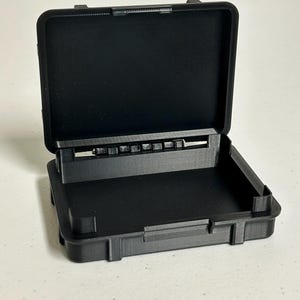 May include: An open, black, rectangular case with a hinged lid. The case is made of a textured material and has a rectangular base. The lid is open, revealing the interior of the case. The case is sitting on a white surface.