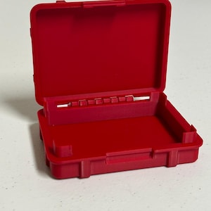 Peut inclure: Mallette en plastique rouge ouverte, de forme rectangulaire. Le boîtier a un couvercle à charnière et un intérieur compartimenté. Le boîtier est vide et repose sur une surface blanche.