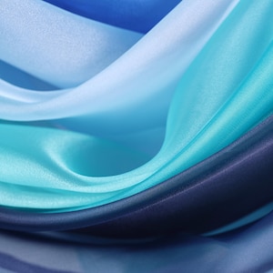 May include: Close-up of flowing fabric in shades of blue. The fabric appears to be silk or satin, with a smooth, lustrous texture. The colors range from light blue to deep navy, creating a gradient effect. The folds and curves of the fabric add depth and dimension.