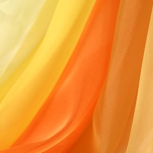 May include: Close-up of flowing fabric in shades of yellow and orange. The sheer material creates a gradient effect, with colors blending from light yellow to a vibrant orange. The fabric is draped, creating soft folds and shadows.