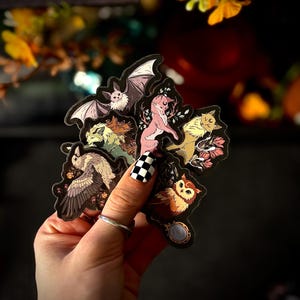 May include: A collection of die-cut stickers featuring various woodland creatures. The stickers include a bat, fox, owl, and other animals, each with unique designs and colors. The stickers are held in a hand with a black and white checkered nail design.