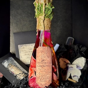 May include: A decorative bottle filled with a pink and red floral arrangement. The bottle is labeled "Herbarium Growth Infusion" and is sealed with a gold-colored wax and green moss. The bottle is in a black box with other items.