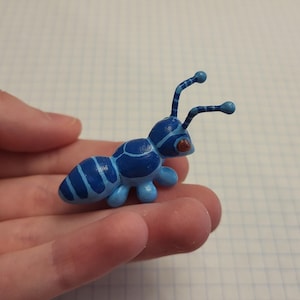 May include: A small, hand-painted figurine of an ant in shades of blue. The insect features a segmented body with light and dark blue stripes, two long antennae with rounded tips, and a single visible reddish-brown eye on the side of its head.