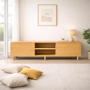May include: A light wood-toned media console with two sliding doors and open shelving. The console has a rectangular shape and sits on short, rounded legs. A lamp, artwork, and decorative items are on top. Three floor pillows in neutral tones are in the foreground.