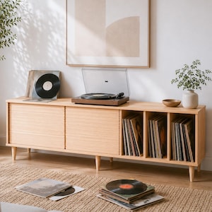 May include: A light wood media console with two sliding tambour doors and four open storage compartments filled with vinyl records. A turntable sits on top, alongside a black vinyl record and a small ceramic vase with a green plant.