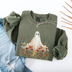 May include: An olive green sweatshirt featuring a whimsical ghost design surrounded by colorful flowers. The ghost has two black eyes and a flowing white sheet. A silver necklace with a circular pendant is draped on the sweatshirt. The sweatshirt is paired with blue jeans.