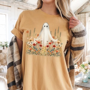 May include: Mustard-colored t-shirt featuring a white ghost design amidst a field of colorful flowers. The ghost has two black eyes. The shirt has short sleeves and a relaxed fit, perfect for casual wear.