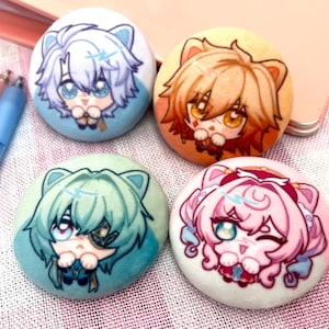 Puede incluir: Cuatro botones redondos y coloridos con ilustraciones de personajes de estilo anime. Los botones presentan personajes con cabello azul, naranja, verde y rosa, cada uno con expresiones faciales y atuendos únicos. Están sobre una superficie con estampado rosa y blanco.