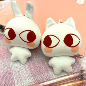 May include: Two white plush toys, one shaped like a rabbit with long ears and the other like a cat with pointed ears. Both have large, red-brown eyes, pink cheeks, and small, star-shaped feet. They are on a pink surface.