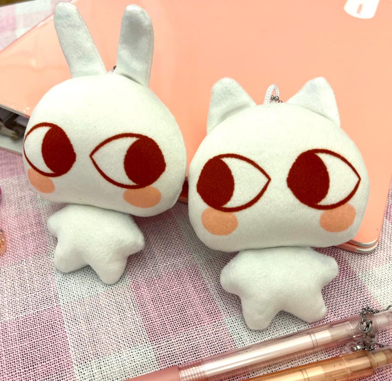 May include: Two white plush toys, one shaped like a rabbit with long ears and the other like a cat with pointed ears. Both have large, red-brown eyes, pink cheeks, and small, star-shaped feet. They are on a pink surface.