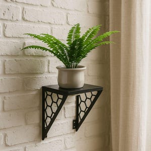 Puede incluir: Una planta de helecho verde en una maceta color crema se encuentra sobre una estantería de pared de metal negro. La estantería tiene un diseño geométrico de panal y está montada en una pared de ladrillo blanco. Una cortina beige está en el fondo.