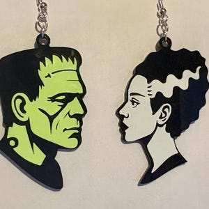 Puede incluir: Pendientes negros y verdes de Frankenstein y la novia de Frankenstein. Los pendientes presentan al icónico monstruo y su novia de perfil, con un contorno negro y detalles verdes y blancos. Los pendientes cuelgan de ganchos plateados.