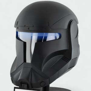 May include: A black Star Wars Republic Commando helmet with a blue visor, displayed on a black stand. The helmet has a matte finish and intricate detailing. A small sign at the base identifies the helmet.