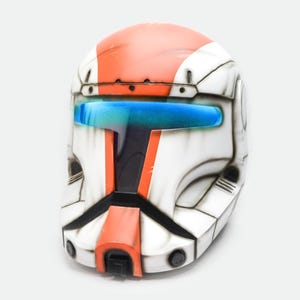 May include: A white and orange helmet with a blue visor. The helmet has black accents and a rounded top. The design is reminiscent of a space trooper's helmet, with a futuristic aesthetic. The helmet is a prop or collectible.