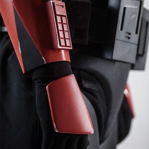 May include: Close-up of a red and black costume. The red armour includes a rectangular panel with a grid of squares and a rectangular plate on the forearm. The costume is worn with black gloves and clothing.