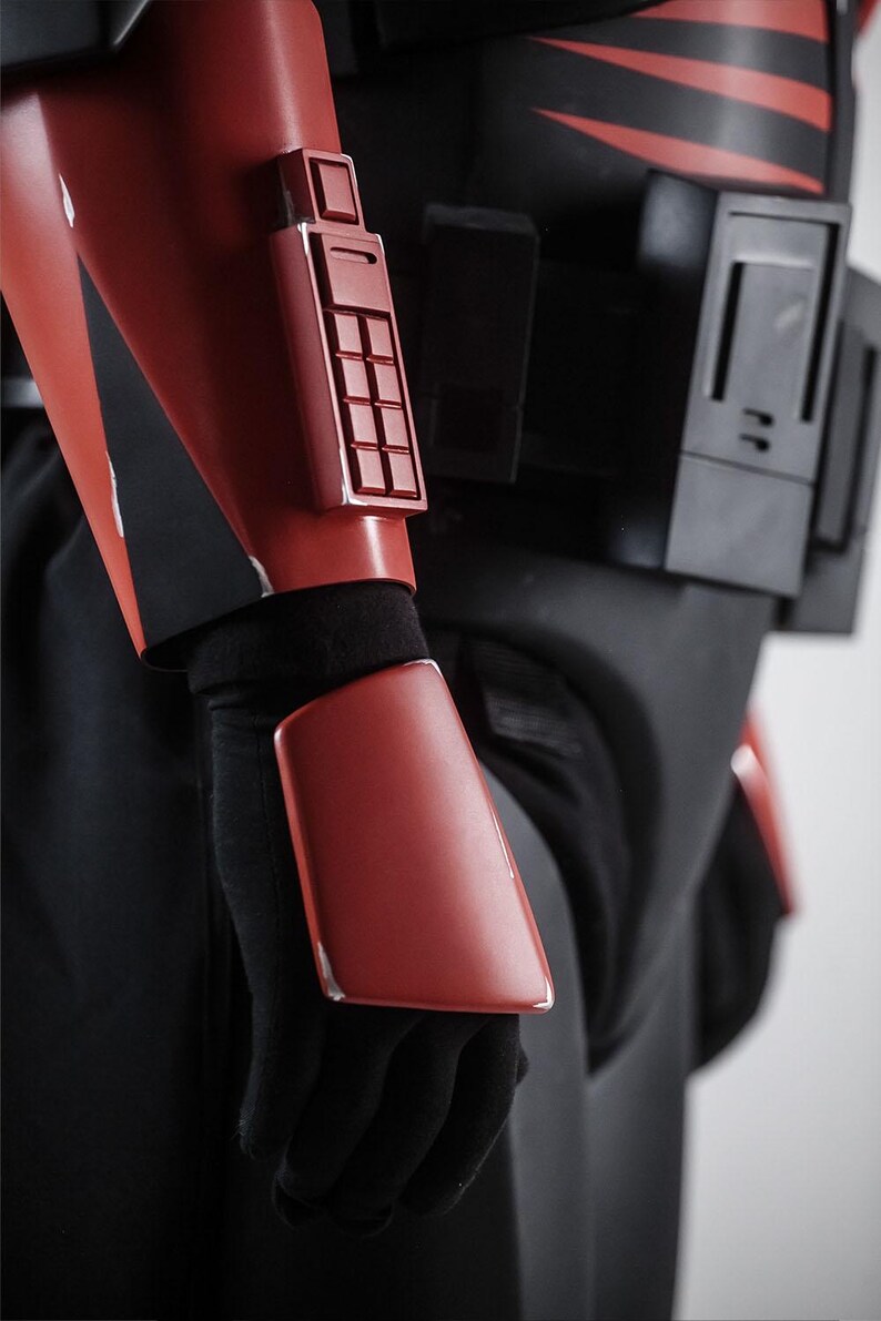 May include: Close-up of a red and black costume. The red armour includes a rectangular panel with a grid of squares and a rectangular plate on the forearm. The costume is worn with black gloves and clothing.