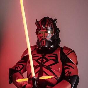 May include: A red and black costume with a yellow double-bladed lightsaber. The costume features a helmet with horns and a black and red design. The lightsaber is held in front of the costume.