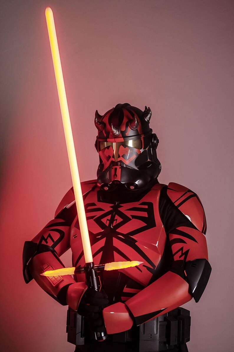 May include: A red and black costume with a yellow double-bladed lightsaber. The costume features a helmet with horns and a black and red design. The lightsaber is held in front of the costume.
