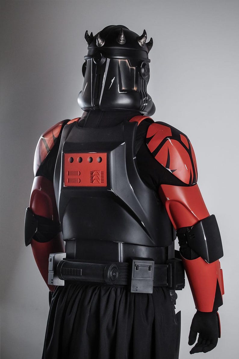 May include: A black and red costume, featuring a helmet with a crown-like design. The armour has red accents on the shoulders and arms, with a black belt and a black skirt. The costume is designed with a futuristic aesthetic.