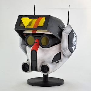 May include: A detailed replica helmet, featuring a black visor with yellow and orange accents, and a white and gray base. The helmet has two black antennas, yellow-tinted goggles, and a red stripe on the front. A black stand supports the helmet.