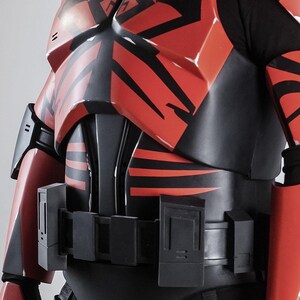 May include: A red and black armoured costume with detailed patterns. The chest and shoulder armour have bold, angular designs. The costume includes a utility belt and black under-layer, creating a striking contrast. The design suggests a futuristic or sci-fi theme.