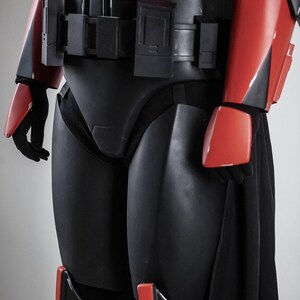 May include: A black and red armoured costume with a sleek design. The costume features red accents on the arms, shoulders, and knees, with a black belt and chest piece. The overall aesthetic suggests a futuristic or science fiction theme.