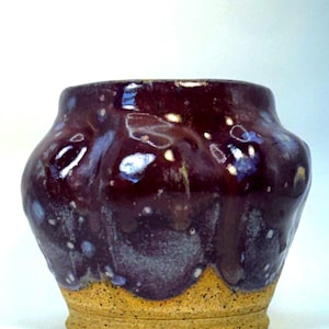 May include: A small ceramic pot with a wide, rounded body and a narrow base. The upper portion features a glossy, deep plum-colored glaze with white speckles and drips, while the lower section has a textured, matte, sandy-beige finish.