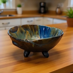 May include: A ceramic bowl with a rounded, organic shape and three short feet. The exterior features a dark, glossy finish, while the interior displays a swirling blend of deep blue, teal, and golden yellow tones with a smooth, reflective glaze.
