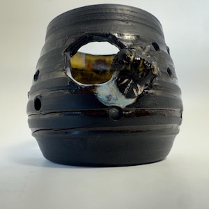 May include: A dark charcoal-colored ceramic vessel with a rounded, tapered shape. The exterior features horizontal ridges, several small circular cutouts, and one larger irregular opening revealing a yellow and white glazed interior surface.