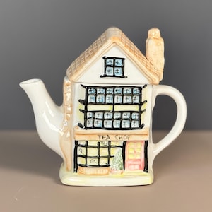 May include: A ceramic teapot shaped like a two-story building with a beige roof and chimney. The teapot is painted with details like windows, a door, and the words "TEA SHOP". The handle and spout are white.