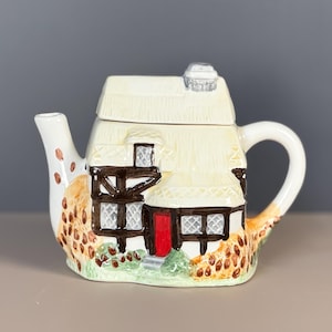 May include: A ceramic teapot shaped like a cottage. The cream-colored roof has a chimney, and the walls are white with brown trim and a red door. The teapot has a curved handle and a spout, with a decorative pattern of brown and orange spots.