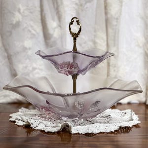 May include: A two-tiered, vintage-style serving dish. The dish features two lavender-colored, scalloped glass bowls connected by a brass-colored metal stand. The bottom bowl has a floral design. The dish is displayed on a white lace doily.