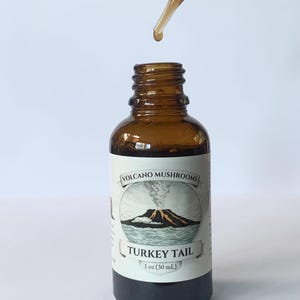 Puede incluir: Una botella de vidrio marrón con una etiqueta blanca que presenta una ilustración de volcán y el texto "TURKEY TAIL". Un cuentagotas dispensa un líquido en la botella. La botella contiene 30 ml de líquido.
