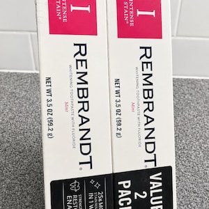 Op de afbeelding: Twee witte rechthoekige dozen Rembrandt Intense Stain whitening tandpasta met fluoride en muntsmaak. Elke doos bevat 99,2 g product. De verpakking heeft een roze koptekst, zwarte tekst en een zwarte basis met 25% meer vlekverwijdering.