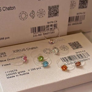 May include: A selection of clear rings, each with a single, coloured gemstone. The gemstones are in shades of pink, green, blue, orange, and clear. The rings are presented on a white surface, with product packaging visible in the background.
