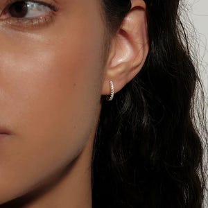 Op de afbeelding: Een close-up van een oor met een kleine, zilveren oorring. De oorring is bezet met kleine, glinsterende steentjes. De persoon heeft donker haar en huid, tegen een effen achtergrond.