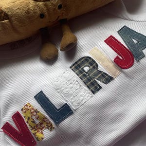 May include: A white, ribbed fabric shirt with the name "VALERIA" appliquéd in various patterned fabrics. A plush, brown baguette-shaped toy with a smiling face rests on top of the shirt.
