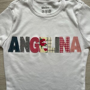 May include: White ribbed t-shirt featuring the name "ANGELINA" appliquéd across the chest. The letters are in different colours and patterns, including floral, plaid, and solid fabrics. The shirt has a crew neck and the brand name "Rita Ora" is visible.