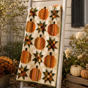 May include: A rectangular quilt featuring a repeating pattern of orange pumpkins and multi-colored star blocks on a cream-colored background. The fabric displays various shades of orange, green, brown, and yellow in a patchwork design with visible stitching.
