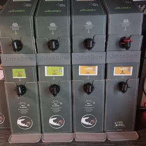 Op de afbeelding: Verschillende JuiceBox-dispensers worden getoond, elk met een donkergrijze buitenkant en een zwarte kraan. De dozen hebben labels met verschillende smaken. De tekst "JuiceBox" is zichtbaar, samen met instructies.