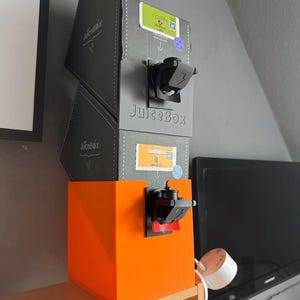 Könnte beinhalten: Ein dreistufiges Saftspendensystem. Die oberen beiden Abschnitte sind grau mit dem Text "JuiceBox" und Etiketten. Der untere Abschnitt ist leuchtend orange. Jeder Abschnitt hat einen schwarzen Ausguss.