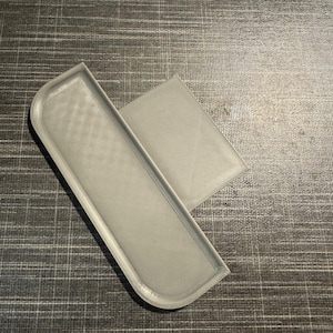 Może przedstawiać: Jasnoszary, prostokątny obiekt drukowany w 3D z zaokrąglonymi narożnikami i podniesioną krawędzią. Osobny, mniejszy, płaski, prostokątny element przylega do większego elementu. Obiekty znajdują się na ciemnoszarej, teksturowanej powierzchni.